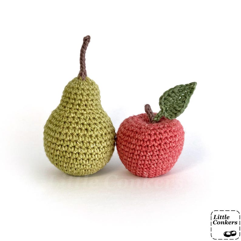 May include: A crocheted pear and apple set. The pear is light green with a brown stem. The apple is red with a brown stem and a single green leaf. The text Little Conkers is visible in the bottom right corner of the image.