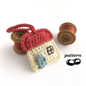 May include: A crocheted house charm with a red roof, white walls, and blue and brown details. The charm is attached to a loop of yarn and is sitting on a spool of red thread.