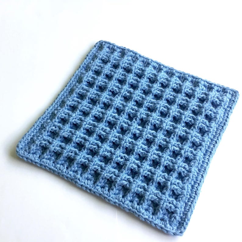 A crocheted dishcloth in blue yarn. The square dishcloth is crocheted in a stitch that gives it a waffle-like appearance and texture.