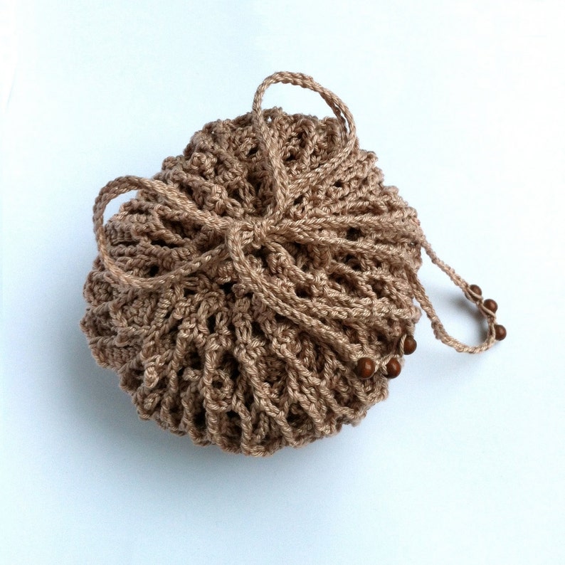 May include: A round, light brown crocheted drawstring pouch featuring an open-work lattice pattern. The bag is cinched at the top with matching crocheted cords that form a bow, and the ends of the cords are decorated with small, round, dark brown wooden beads.