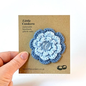 May include: A blue and light blue crocheted flower with a light blue button in the center. The flower is on a brown card with the text "Little Conkers, sustainable handmade gifts & crafts, www.littleconkers.co.uk".