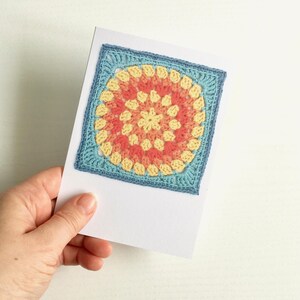 May include: A white greeting card featuring a square crochet motif centered on the front. The crochet design is a circular pattern in shades of pale yellow, coral, and orange, framed by a light blue border that squares off the circular shape.