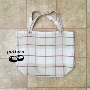 Puede incluir: Una bolsa de tela blanca con un patrón de cuadros rojos y blancos. La bolsa tiene dos asas y está hecha de tela tejida.