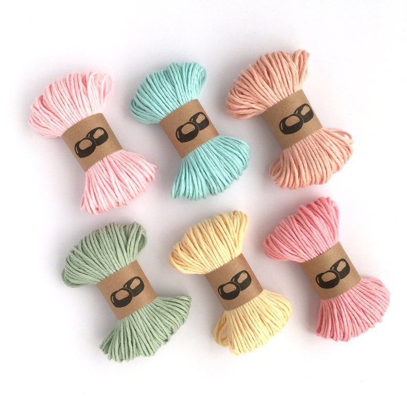 May include: Six bundles of twisted cord arranged in two rows. The colors include light pink, mint green, peach, sage green, pale yellow, and rose pink. Each bundle is secured with a brown paper band featuring a black icon of two rounded shapes.