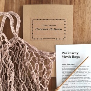 May include: A light pink crocheted mesh bag with two handles, a square brown envelope labeled Little Conkers Crochet Pattern, and a white instruction sheet titled Packaway Mesh Bags, accompanied by a wooden crochet hook for crafting.