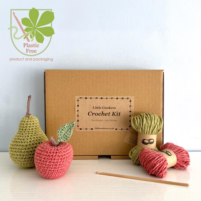 May include: A Little Conkers Crochet Kit in a brown cardboard box with a label, featuring a crocheted green pear, a red apple with a green leaf, two skeins of yarn in green and red, and a wooden crochet hook, with a plastic-free logo.