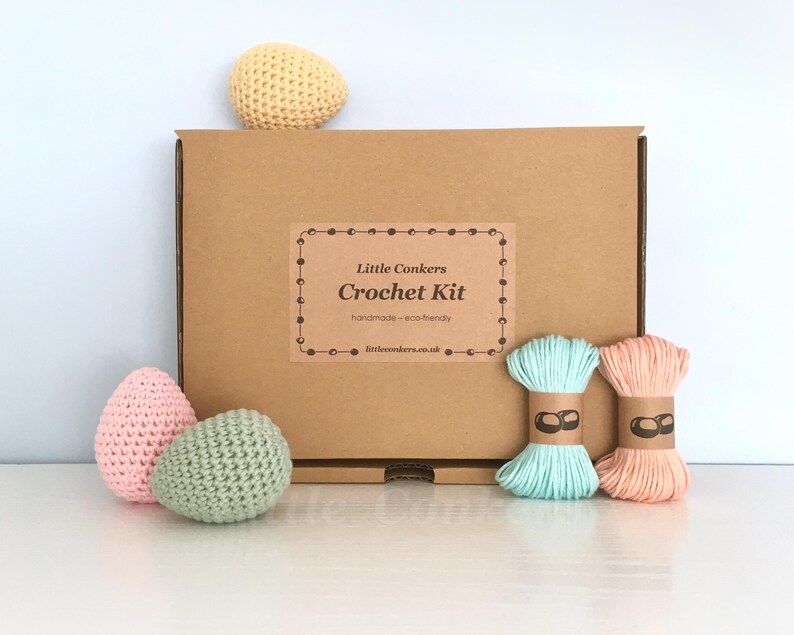 May include: A brown cardboard box labeled Little Conkers Crochet Kit with handmade eco-friendly text. The kit includes a yellow crocheted egg, a pink crocheted egg, a green crocheted egg, and two skeins of yarn in light blue and peach colors.