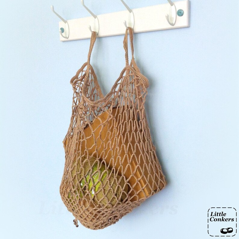 May include: A light brown crocheted net bag with an open mesh design and two handles, hanging from a white wall-mounted hook rack. Inside the bag, a round ball of green yarn and a rectangular brown object are visible. The text Little Conkers is present.