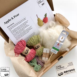 May include: A craft kit in a cardboard box containing a printed instruction sheet titled Apple & Pear, a 3.50 mm bamboo crochet hook, a small sewing needle, white stuffing material, and four skeins of yarn in red, light green, dark green, and purple.