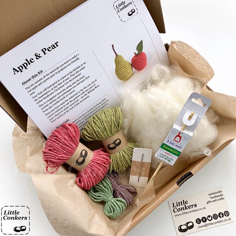 May include: A craft kit in a cardboard box containing a printed instruction sheet titled Apple & Pear, a 3.50 mm bamboo crochet hook, a small sewing needle, white stuffing material, and four skeins of yarn in red, light green, dark green, and purple.