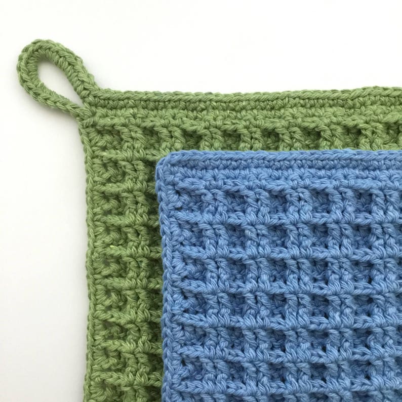 Two crocheted dishcloths, one in blue yarn, one in green yarn. The dishcloths are crocheted with a stitch that gives them a waffle-like appearance and texture. There is a hanging loop in one corner of the dishcloths.