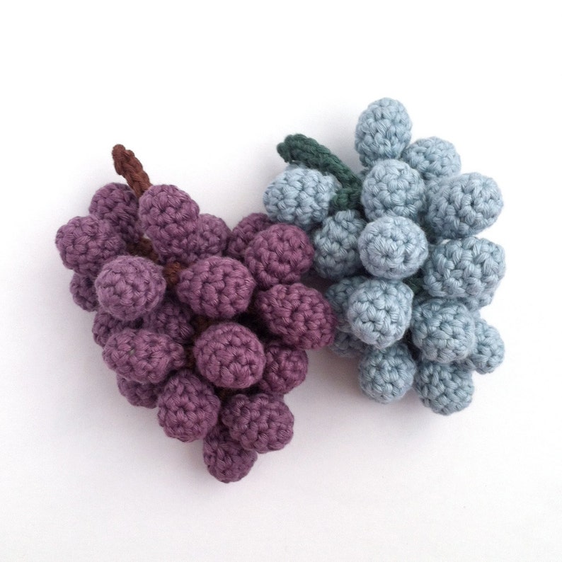 Two crocheted bunches of grapes. One is a realistic bunch of red grapes in purple and brown yarn. The other is made of pale blue grapes.
