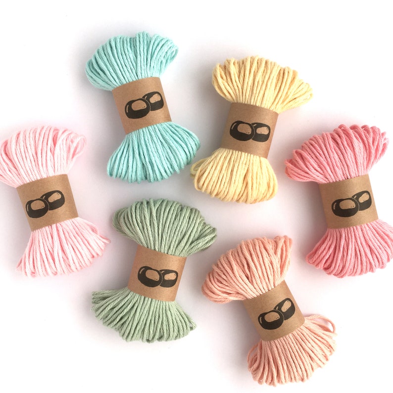 May include: Six bundles of cotton cord in pastel colors including light blue, yellow, light pink, sage green, peach, and coral. Each bundle is wrapped in a brown paper band featuring a black graphic logo of two overlapping oval shapes.