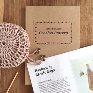 May include: A crochet pattern kit for packaway mesh bags, featuring a dusty pink crocheted sphere, a wooden crochet hook, and a printed instruction booklet with the text Little Conkers Crochet Pattern and Packaway Mesh Bags.