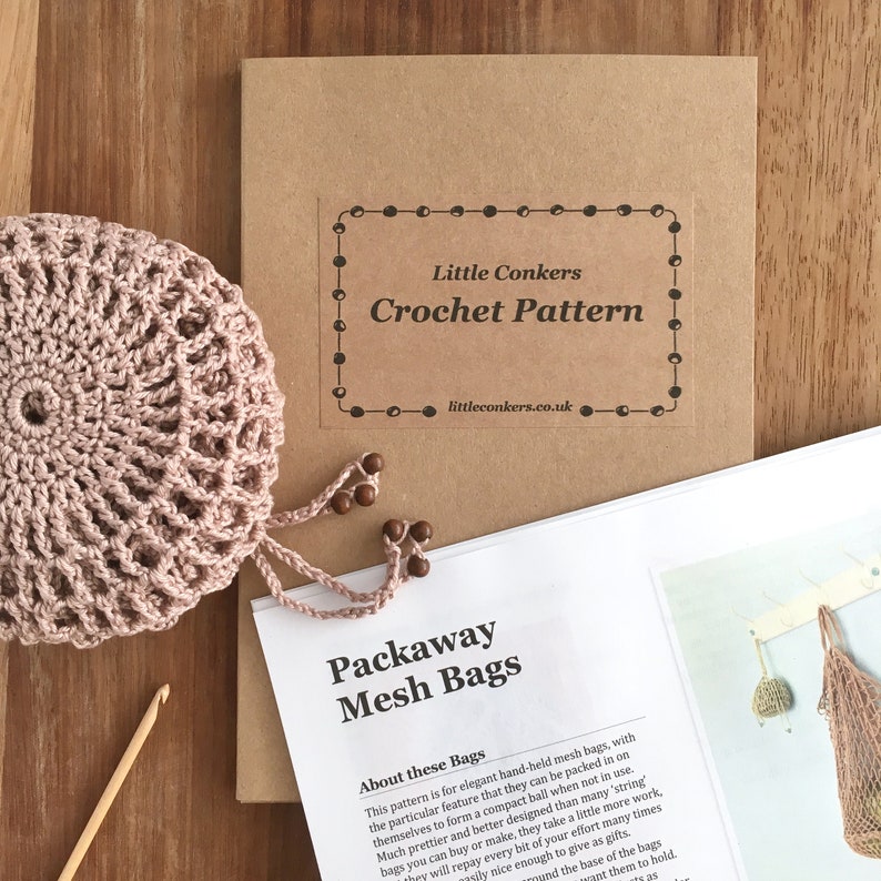 May include: A crochet pattern kit for packaway mesh bags, featuring a dusty pink crocheted sphere, a wooden crochet hook, and a printed instruction booklet with the text Little Conkers Crochet Pattern and Packaway Mesh Bags.