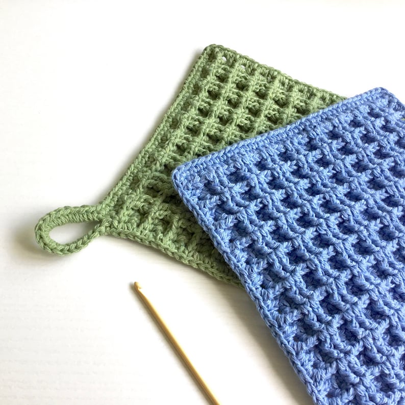 Two crocheted dishcloths, one in blue yarn, one in green yarn. The square dishcloths are crocheted with a stitch that gives them a waffle-like appearance and texture. There is a hanging loop in one corner of the dishcloths.