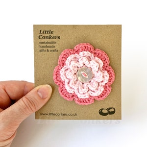May include: A pink and white crocheted flower with a silver button in the center. The flower is on a brown card with the text "Little Conkers, sustainable handmade gifts & crafts, www.littleconkers.co.uk".
