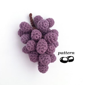 Puede incluir: Un racimo de uvas de color morado tejido a crochet con un tallo marrón. La palabra "pattern" está escrita en texto negro con dos círculos negros debajo.