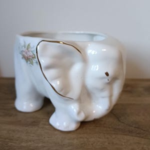 May include: A white ceramic planter shaped like an elephant with a gold-trimmed ear and a small floral decal on the side. The elephant figure features a hollow opening at the top for plants, with a glossy finish and a trunk curled downward.