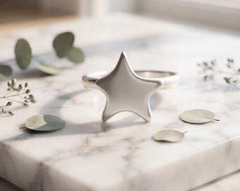 May include: A silver-toned ring featuring a prominent five-pointed star design on the front. The star has smooth, rounded edges and a polished metallic finish. The band of the ring is simple and circular, connecting to the back of the star.