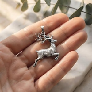 May include: A silver-toned metal pendant shaped like a leaping stag with detailed antlers and a textured body. The pendant features a small loop at the top for attachment to a chain, showcasing a metallic finish with dark recessed areas for contrast.