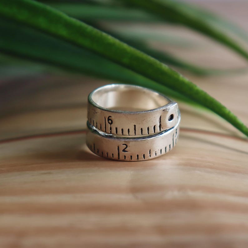 May include: A silver ring designed to resemble a flexible measuring tape. The band features engraved black markings, including the numbers 2 and 6, mimicking the appearance of a standard imperial measuring tape with visible hash marks.