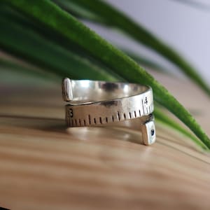 May include: A silver-toned metal ring designed to resemble a flexible measuring tape. The band features engraved black hash marks and the numbers 3 and 4, with a small dark circular accent at one of the open, slightly flared ends.