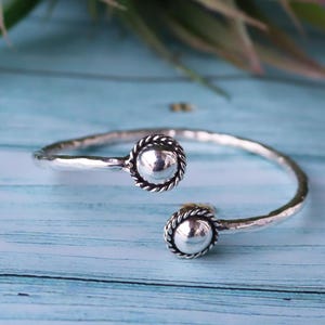 May include: A silver-toned open cuff bracelet featuring a hammered metal band. The two ends of the bracelet are finished with smooth, polished spherical beads, each encircled by a decorative twisted rope-style metal border detail.
