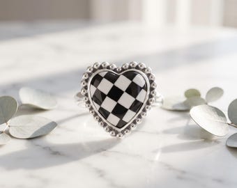 May include: A silver ring featuring a heart-shaped centerpiece with a black and white checkered pattern. The heart is framed by a border of small, raised silver beads, and the ring band is made of polished silver metal.