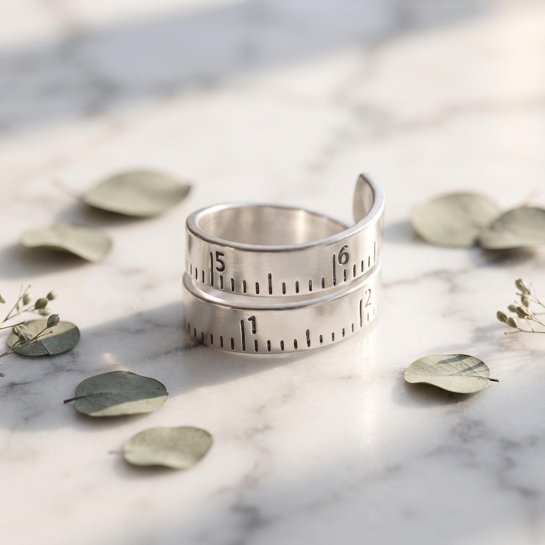 May include: A silver-toned wrap ring featuring a design that mimics a measuring tape. The band displays engraved black markings and numbers, including the digits 1, 5, and 6, arranged in a continuous spiral pattern around the finger band.