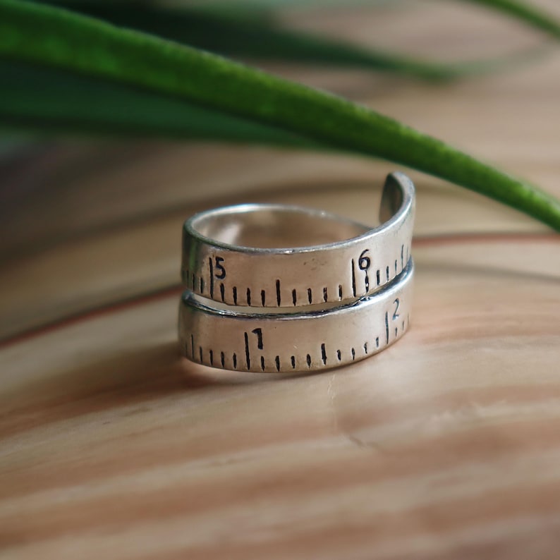 May include: A silver-toned metal ring designed to resemble a coiled measuring tape. The band features black engraved markings and numbers, including the digits 1, 2, 5, and 6, mimicking the appearance of a ruler wrapped around the finger.