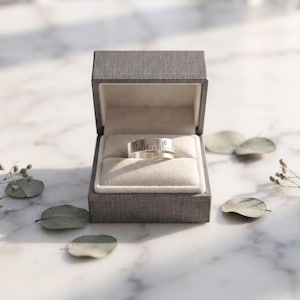 May include: A silver-toned metal ring featuring a wrap-around design with engraved black markings resembling a ruler, including the numbers 15 and 16, presented inside a dark gray textured jewelry box with a cream-colored velvet interior.