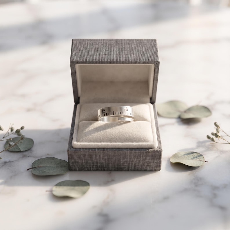 May include: A silver-toned metal ring featuring a wrap-around design with engraved black markings resembling a ruler, including the numbers 15 and 16, presented inside a dark gray textured jewelry box with a cream-colored velvet interior.
