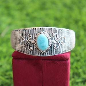May include: A silver-toned cuff bracelet featuring a central oval-shaped light blue stone. The metal surface has a textured, stippled finish and is decorated with symmetrical, swirling filigree patterns on both sides of the stone setting.