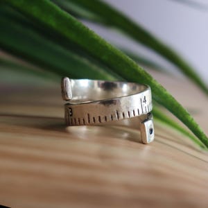 May include: A silver-toned metal ring designed to resemble a flexible measuring tape. The band features engraved black hash marks and the numbers 3 and 4, with a small dark circular accent at one of the open, slightly flared ends.