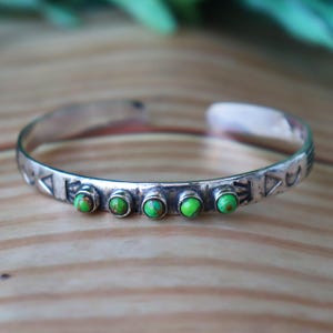 May include: A silver cuff bracelet featuring five small, round, bright green stones set in a row along the centre. The metal band is engraved with geometric patterns, including triangles and crescent shapes, along the outer surface.