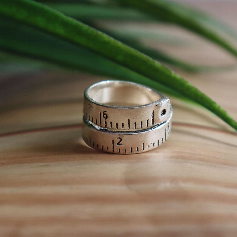 May include: A silver ring designed to resemble a flexible measuring tape. The band features engraved black markings, including the numbers 2 and 6, mimicking the appearance of a standard imperial measuring tape with visible hash marks.