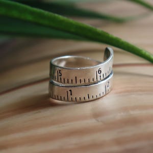 May include: A silver-toned metal ring designed to resemble a coiled measuring tape. The band features black engraved markings and numbers, including the digits 1, 2, 5, and 6, mimicking the appearance of a ruler wrapped around the finger.