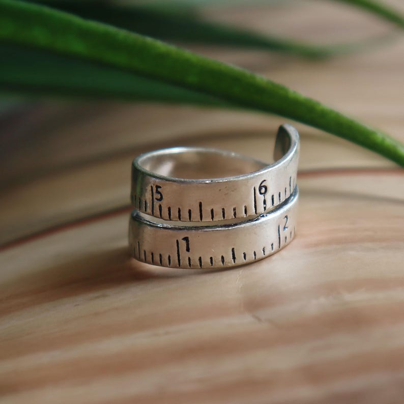 May include: A silver-toned metal ring designed to resemble a coiled measuring tape. The band features black engraved markings and numbers, including the digits 1, 2, 5, and 6, mimicking the appearance of a ruler wrapped around the finger.