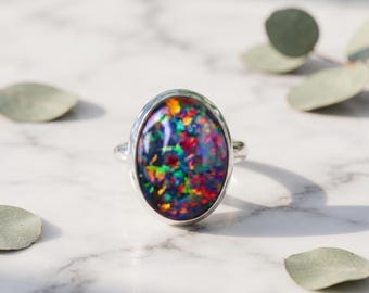 May include: A silver ring featuring a large, oval-shaped cabochon stone with a dark base color and vibrant, multi-colored iridescent flecks of red, green, blue, and yellow throughout the surface, set in a smooth, polished metal bezel.