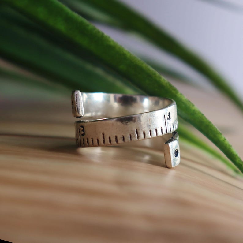 May include: A silver-toned metal ring designed to resemble a flexible measuring tape. The band features engraved black hash marks and the numbers 3 and 4, with a small dark circular accent at one of the open, slightly flared ends.