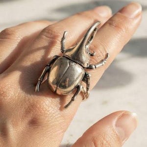 May include: A silver-toned ring featuring a large, detailed rhinoceros beetle design. The beetle has a prominent horn, segmented legs, and a smooth, metallic finish. The ring is worn on a finger, showcasing the intricate, realistic anatomy of the insect.