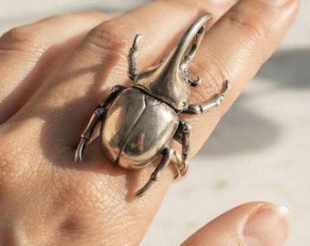 May include: A silver-toned ring featuring a large, detailed rhinoceros beetle design. The beetle has a prominent horn, segmented legs, and a smooth, metallic finish. The ring is worn on a finger, showcasing the intricate, realistic anatomy of the insect.