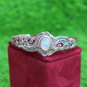 May include: A silver cuff bracelet featuring an oval-shaped, iridescent moonstone set in the center. The metal band displays intricate, raised scrollwork and rope-like detailing surrounding the stone, with a polished finish throughout.