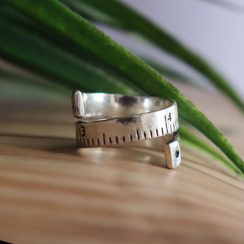 May include: A silver-toned wrap ring designed to resemble a flexible measuring tape. The band features engraved black markings and numbers, including the visible digits 3 and 4, with a small dark accent at one of the tapered ends of the ring.