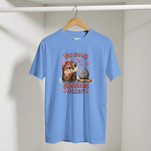 Könnte beinhalten: Ein hellblaues T-Shirt mit einer Cartoon-Illustration eines braunen Otters mit einer rosa gepunkteten Schleife, der einen grauen Stein betrachtet. Der Text "You are my significant otter!" ist von kleinen rosa Herzen auf der Vorderseite umgeben.