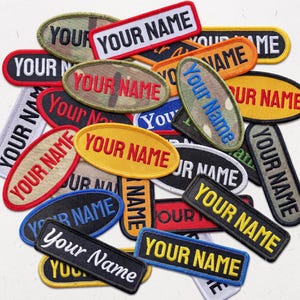 Könnte beinhalten: Eine Sammlung von rechteckigen und ovalen gestickten Aufnähern mit dem Text YOUR NAME in verschiedenen Schriftarten, Farben und Hintergrundmustern, darunter Schwarz, Gelb, Rot, Blau, Beige und Tarnfarben, mit gestickten Rändern.