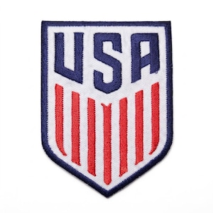 May include: A shield-shaped embroidered patch featuring the text USA in bold navy blue letters at the top, positioned above seven vertical red stripes on a white background, all enclosed within a thick navy blue border with a pointed bottom edge.
