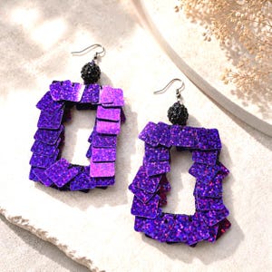 Könnte beinhalten: Ein Paar rechteckige Ohrhänger mit Schichten aus schimmernden violetten quadratischen Pailletten. Jeder Ohrring ist mit einer kleinen, texturierten schwarzen Kugelperle und einem silberfarbenen Metallhaken für Ohrlöcher versehen.