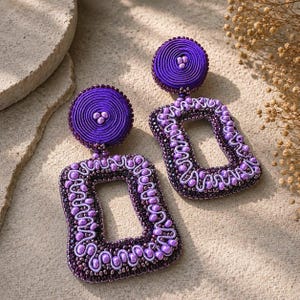 May include: A pair of purple dangle earrings featuring a circular top component made of coiled thread and a rectangular lower section decorated with intricate beadwork, metallic seed beads, and wavy light purple cord detailing throughout.